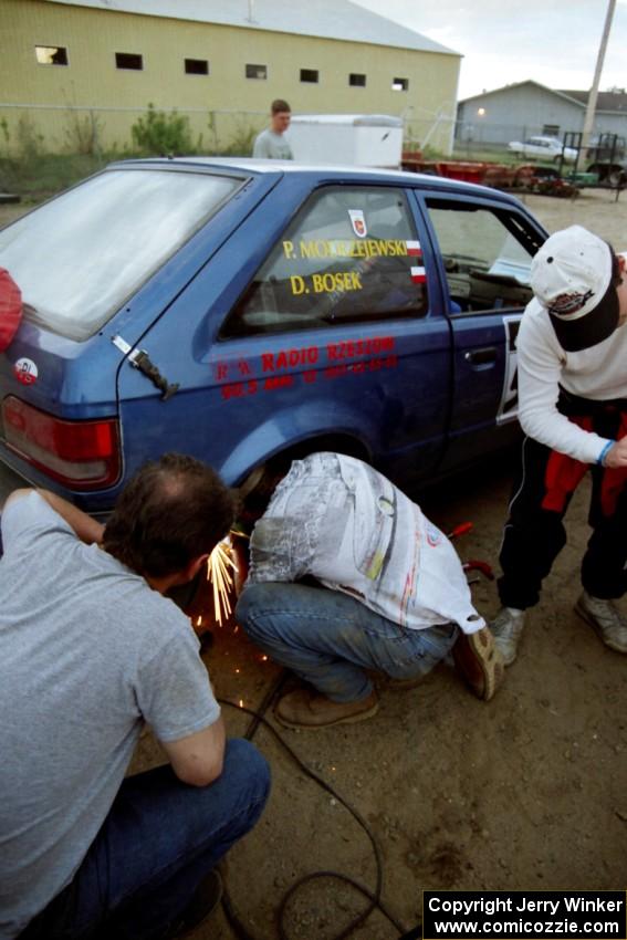 The Darek Bosek / Piotr Modrzejewski Mazda 323GTX receives repairs at service after driving with a flat on a stage.