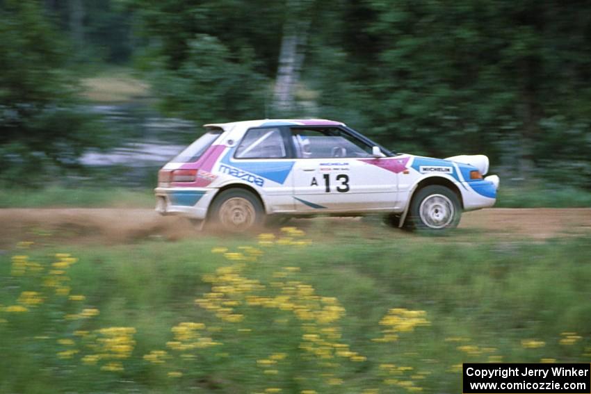 The Peter Moodie / Mike Fennell Mazda 323GTR powers away from spectators at the county road.