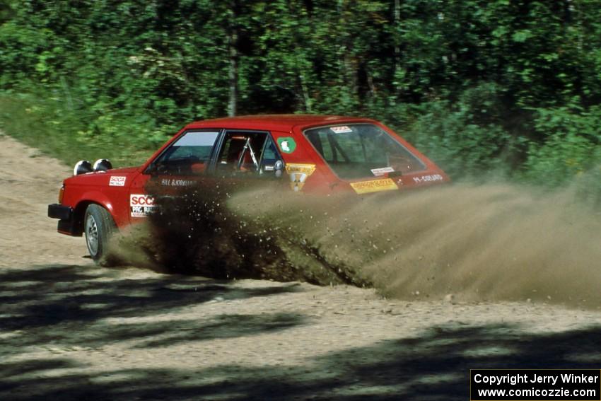 Bill Bjorklund / Al Kintigh kick up a spray of gravel at the crossroads in their Dodge Omni.